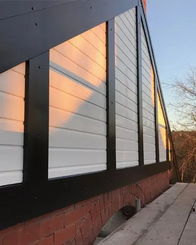 Building wall with black framing and white paneling, sunlight reflecting off the surface. Brick foundation visible below.