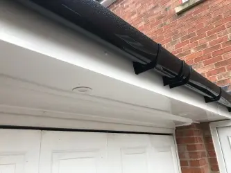Black gutter attached to a white overhang above a garage door with a brick wall in the background.
