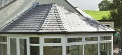 House with a slate roof and large windows, surrounded by greenery.