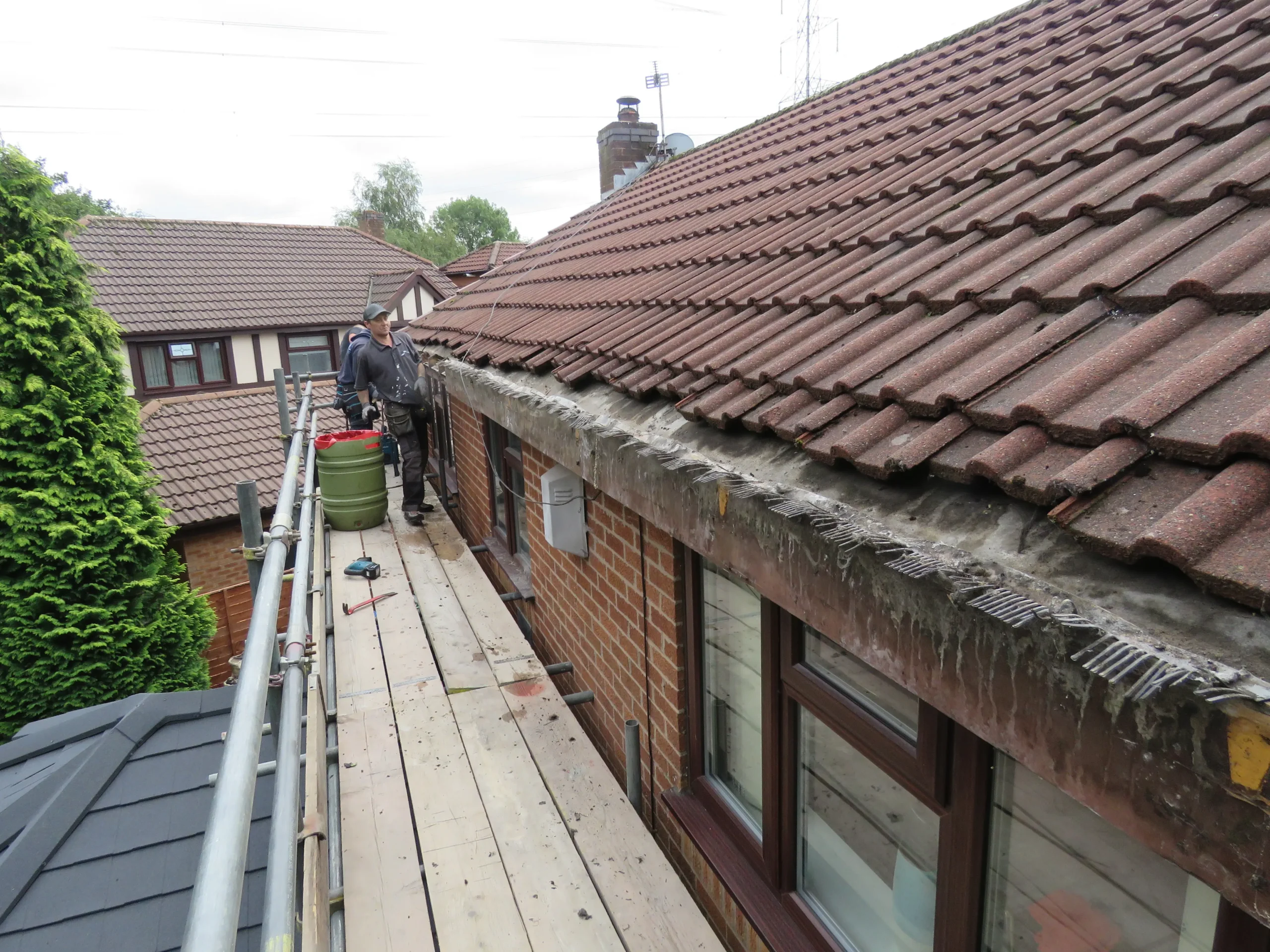 Two workers on scaffolding cleaning a roof, with a green bucket and tools visible nearby.