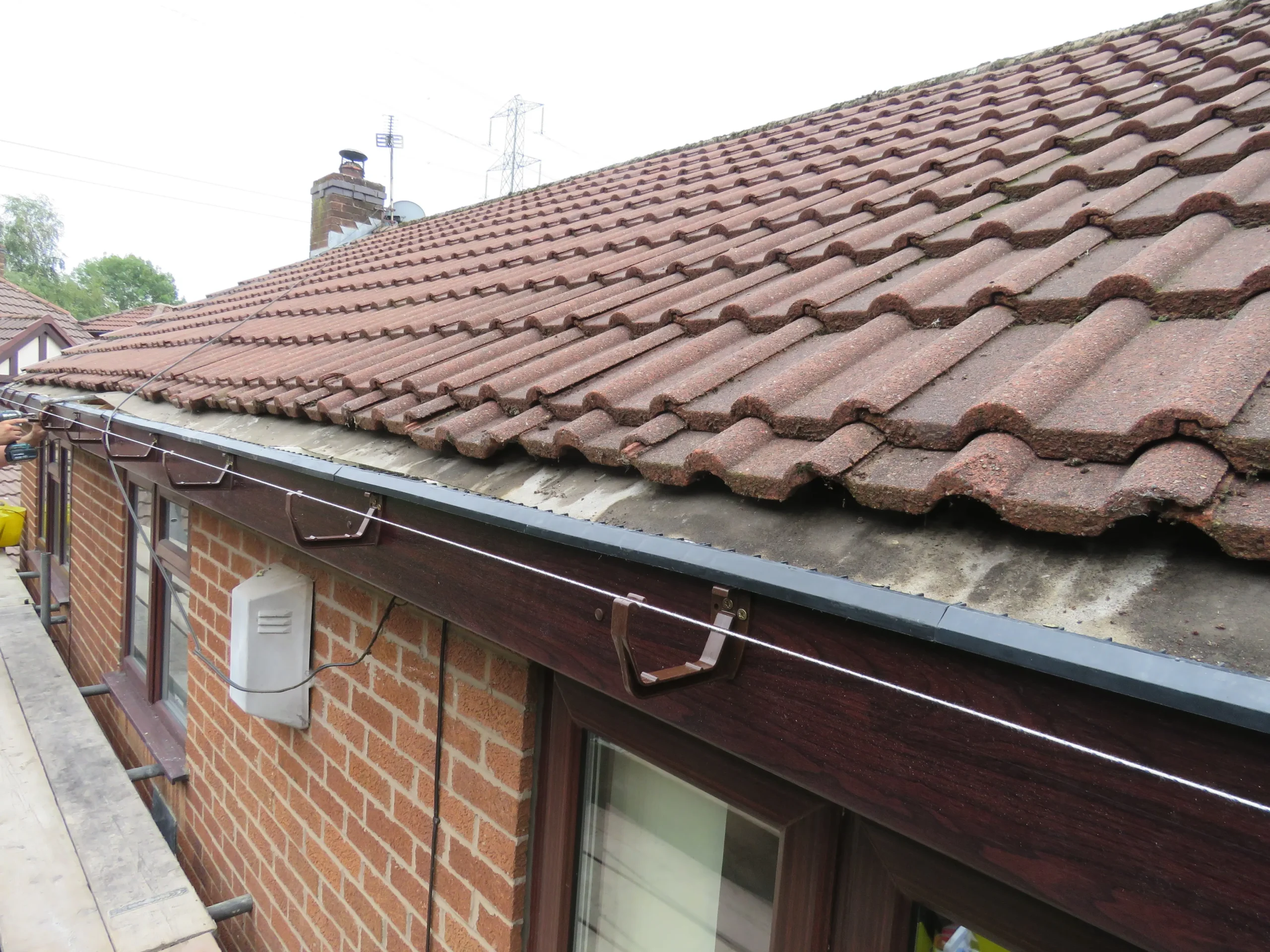 Roof with red tiles, guttering, and a white electricity box on a brick wall, scaffolding partially visible.