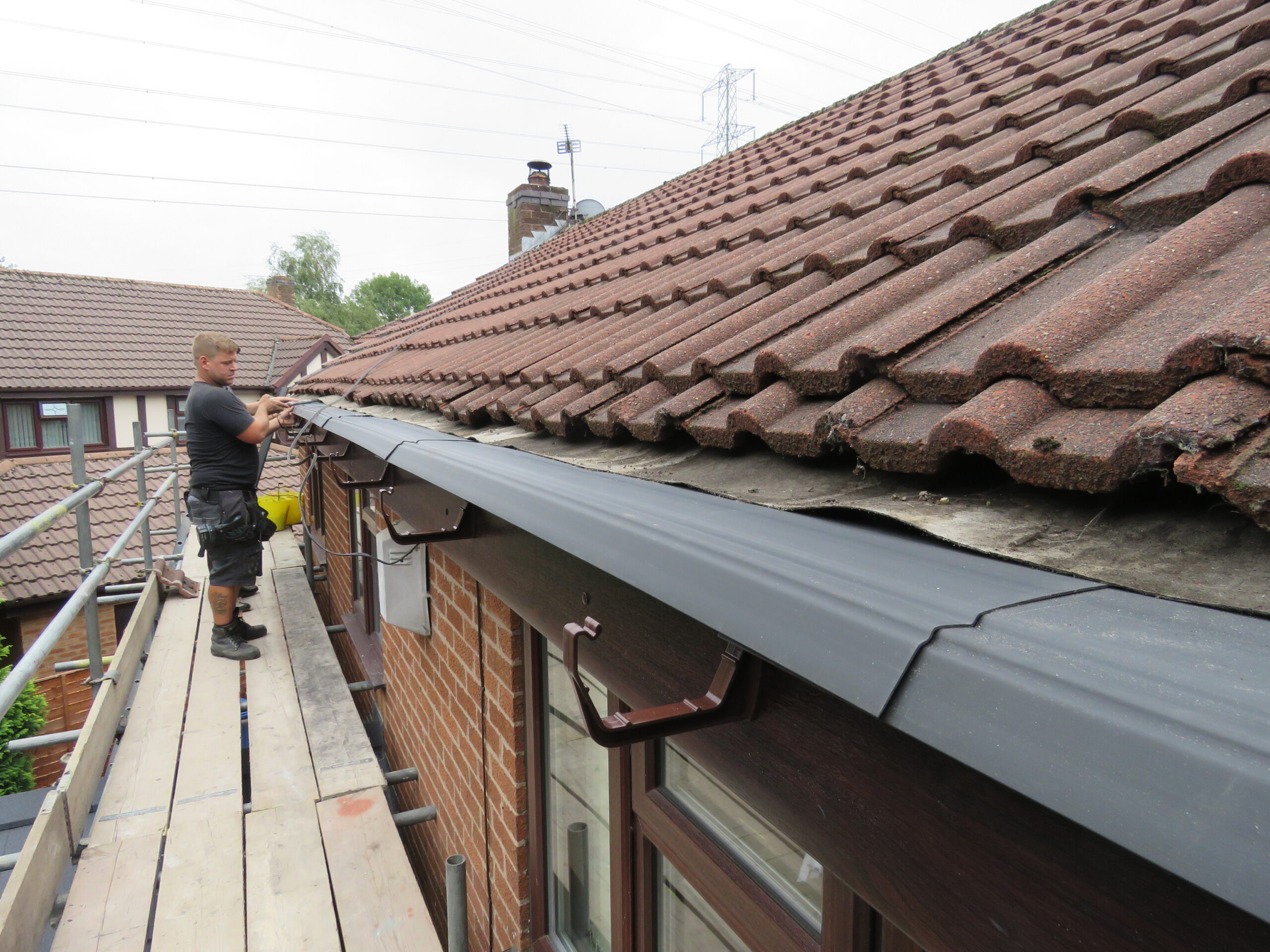 Worker installing new gutter on a house roof while standing on scaffolding.