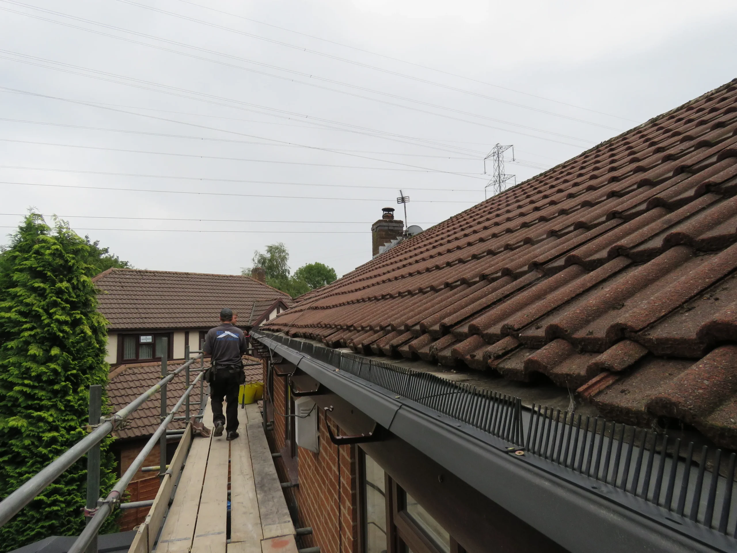 Worker walking on scaffolding beside a brown tiled roof with overcast sky and power lines in the background.