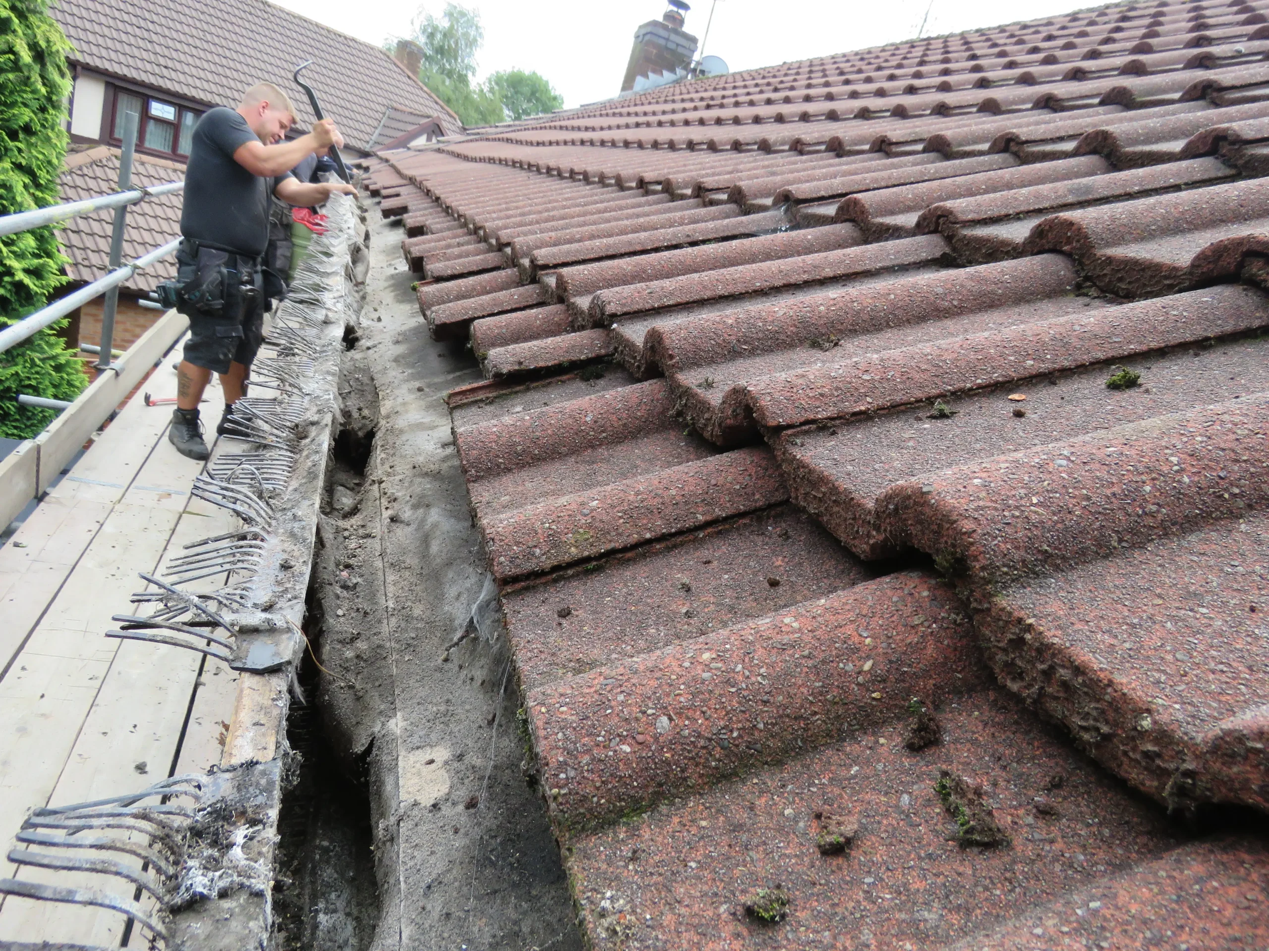 Worker repairing the edge of a roof with a tool, surrounded by roofing tiles and a scaffold.