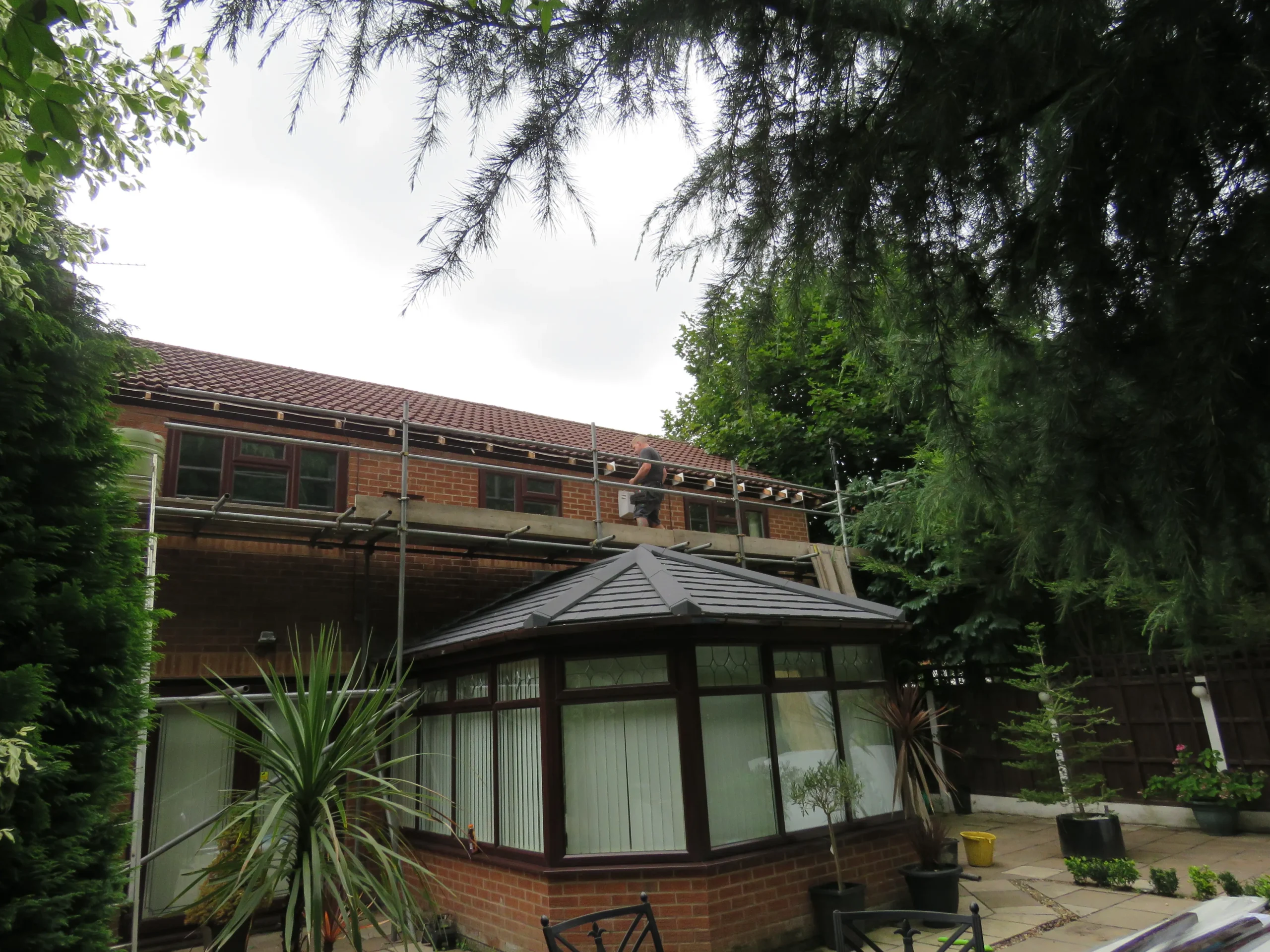 Builder working on roof with scaffolding above a sunroom in a residential garden.