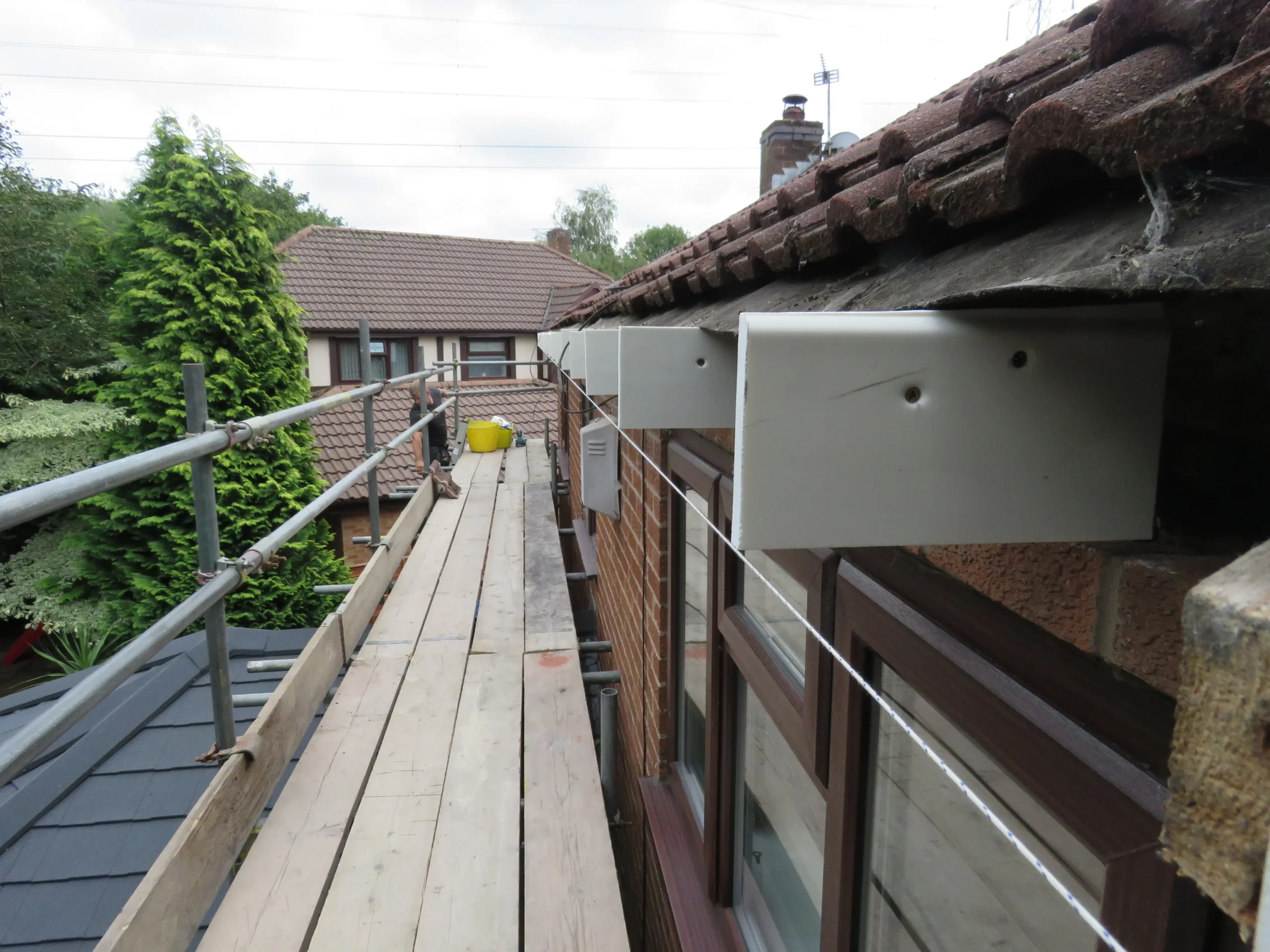 Scaffolding along a wooden walkway beside a building with tiled roof and adjoining trees.