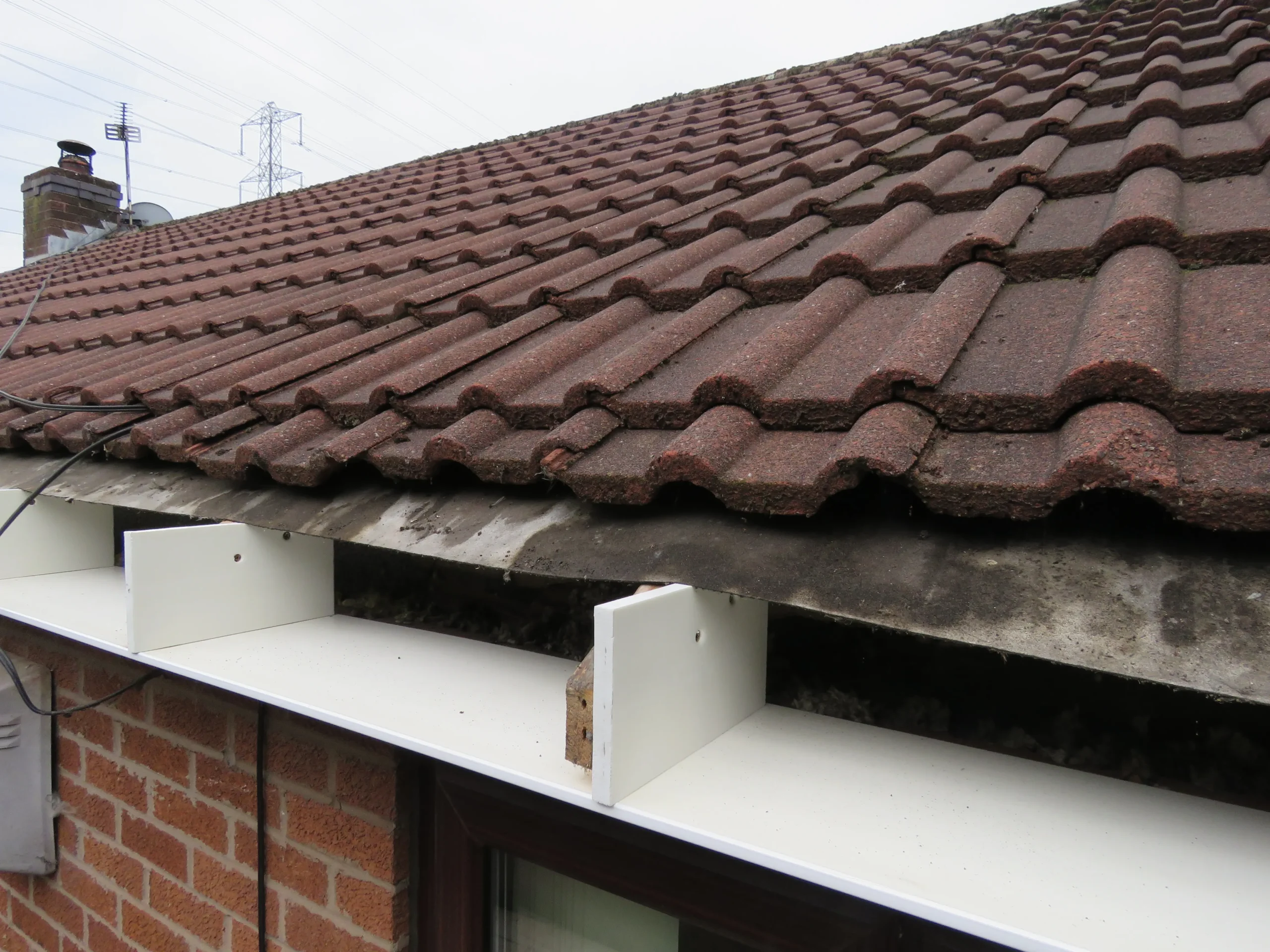 Brown tiled roof with white guttering and visible dirt along the edge, with electrical wires and chimney in background.