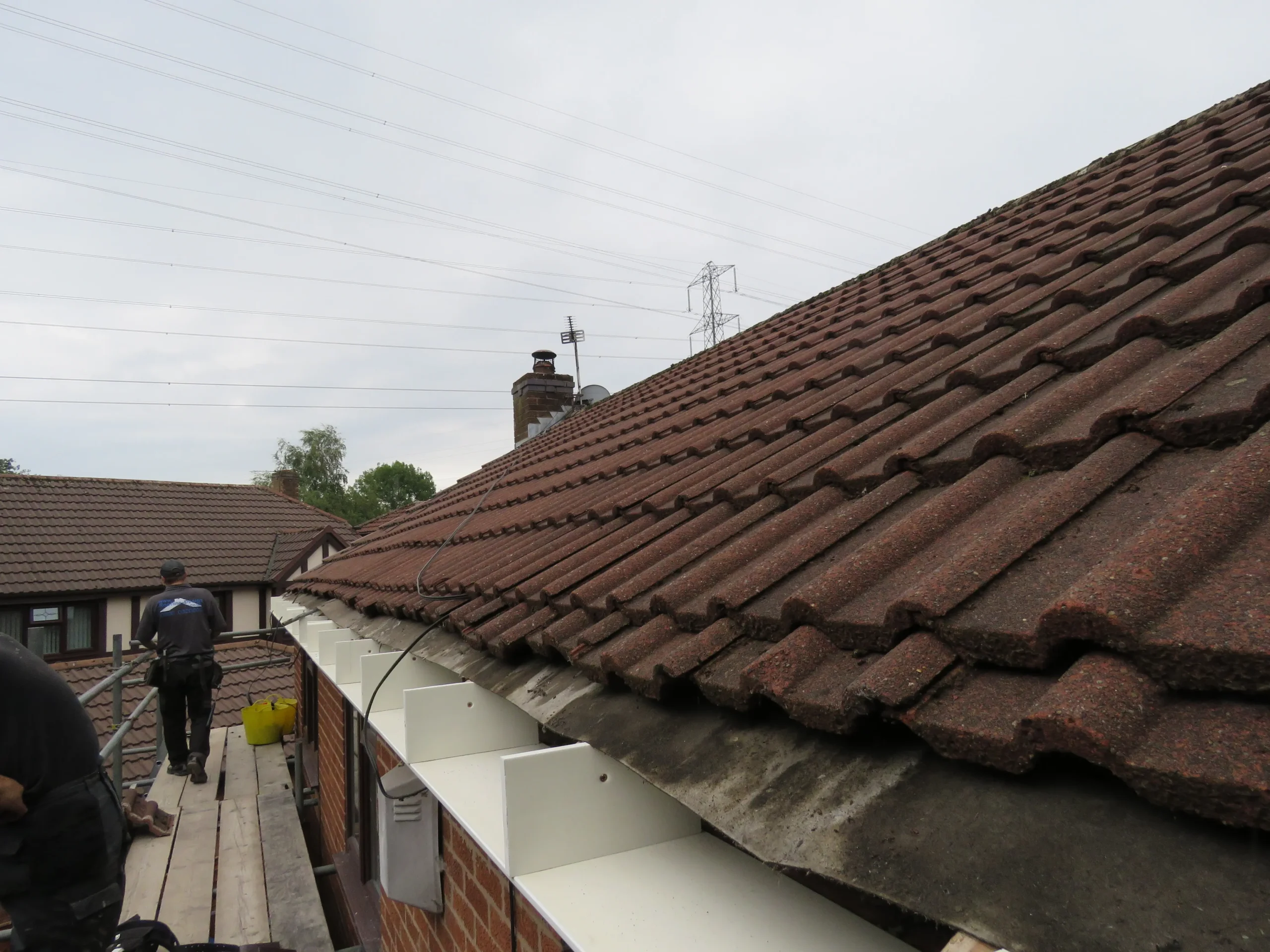 Rooftop with reddish-brown tiles, worker on scaffolding, and cloudy sky with power lines in the background.
