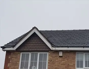 Roof of a house with slate shingles and brick walls, featuring a triangular gable and windows below.