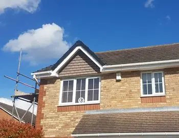 Two-story brick house with a peaked roof, scaffolding on the left, and blue sky with clouds in the background.