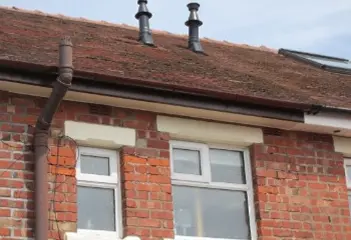 Brick house exterior with two chimneys on the roof and three windows visible on the first floor.