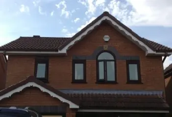 Two-story brick house with a sloped roof, arched window, and dark-colored framed windows under a partly cloudy sky.