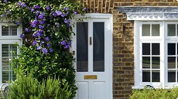 White door surrounded by purple flowers and brick wall, with two windows on either side.