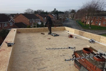 Construction worker sweeping a newly installed flat roof, tools and houses in the background.