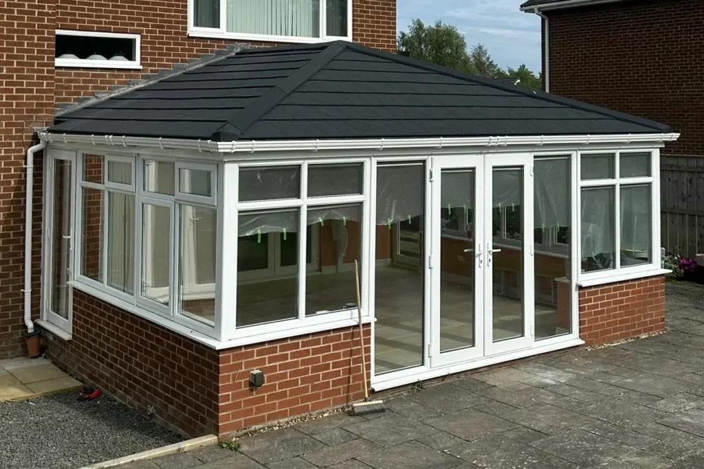 Conservatory with white frames, glass walls, and a black roof, alongside a brick house and stone patio.
