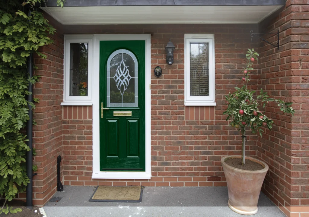 Green front door with decorative glass panel, flanked by two windows and a potted tree, on brick wall.