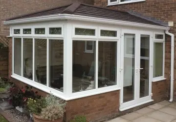White conservatory with multiple windows, a door, and a tiled roof, surrounded by a patio and garden area.
