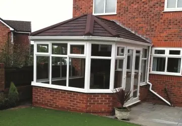 Conservatory with a brown roof and white frames, surrounded by grass and a brick house exterior.