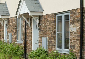 Row of stone house exteriors with grey shutters, white doors, and green grass in the foreground.