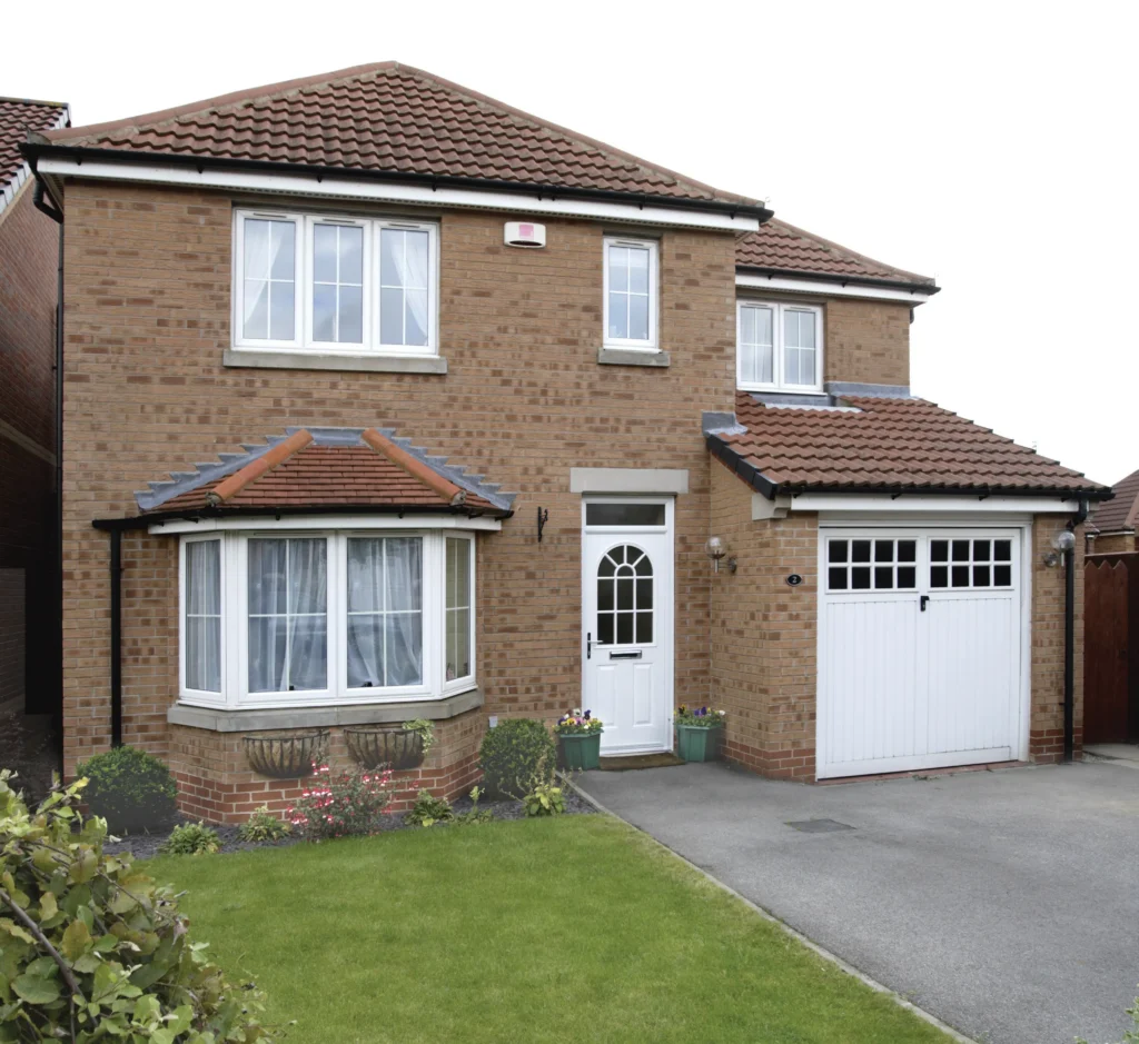 Two-story brick house with a white front door, garage, and bay window, surrounded by grass and flower pots.