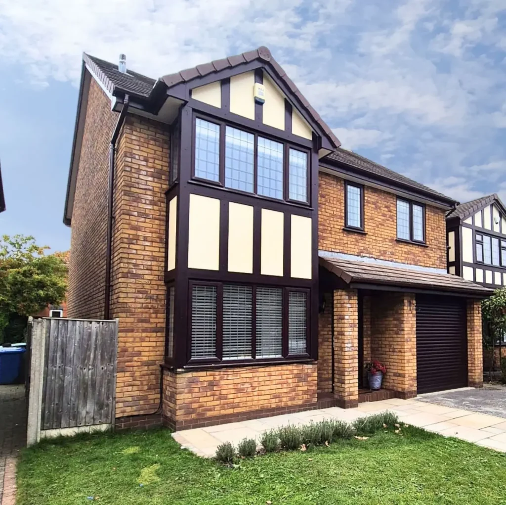 Two-story brick house with cream and brown accents, front garden, and paved walkway under a blue sky.