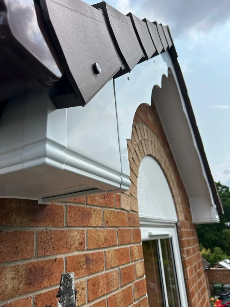 Roofline with brown tiles and white guttering against a brick wall and cloudy sky.