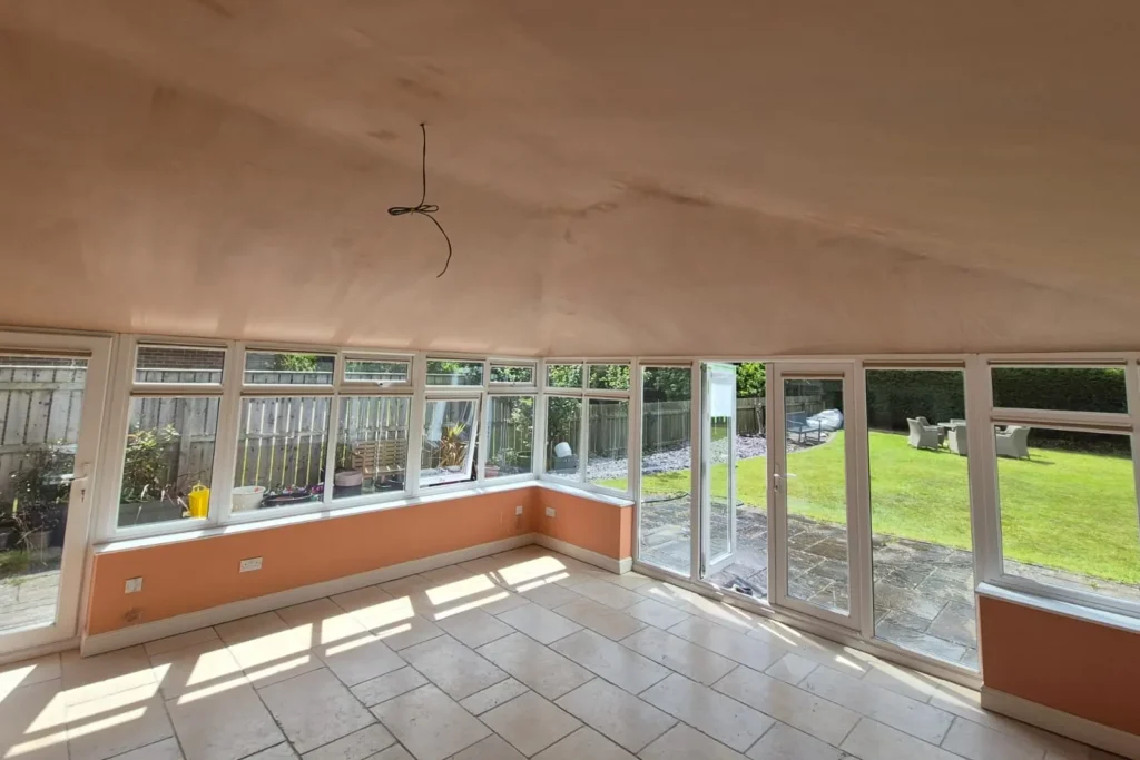 Sunroom with large windows, tiled floor, and an open door leading to a green garden area with patio furniture.